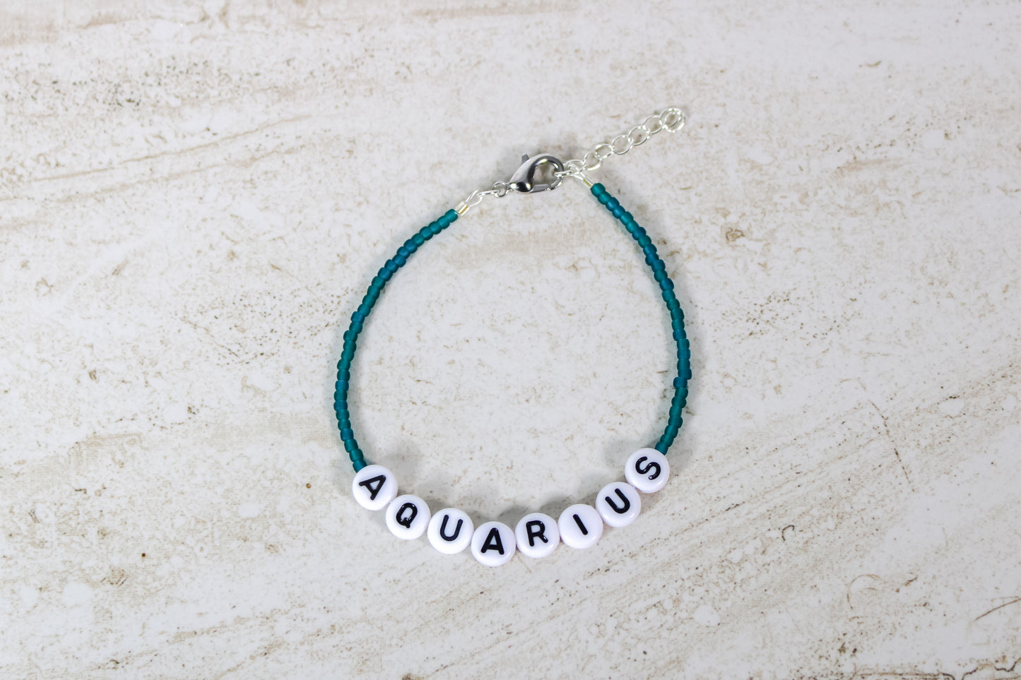 A teal colored seed bead bracelet with a Aquarius in round letter beads and silver lobster clasp and extender chain.