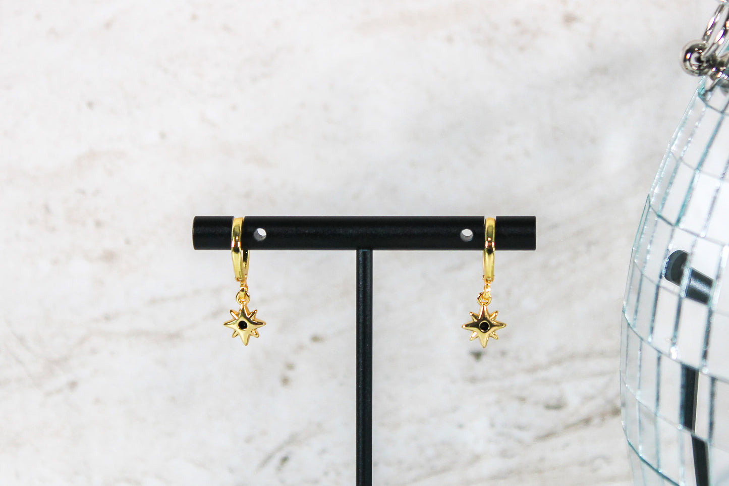 A pair of gold huggie hoop earrings with small black cubic zirconia north star charms are displayed on a t bar, showing the back of the earrings.