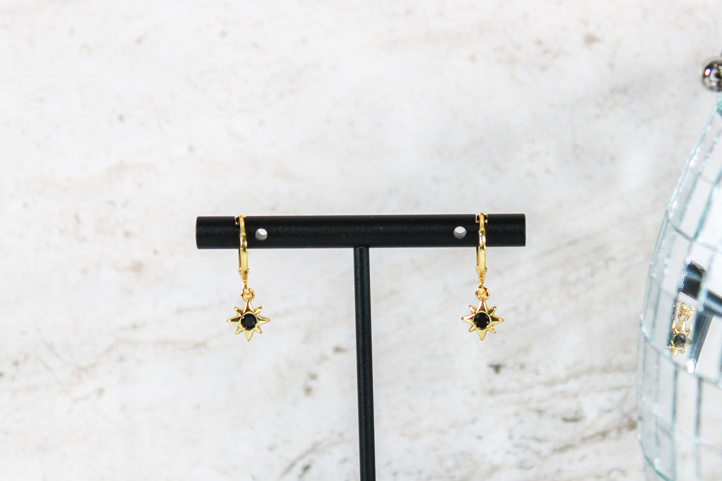 A pair of gold huggie hoop earrings with small blue cubic zirconia north star charms are displayed on a t bar.