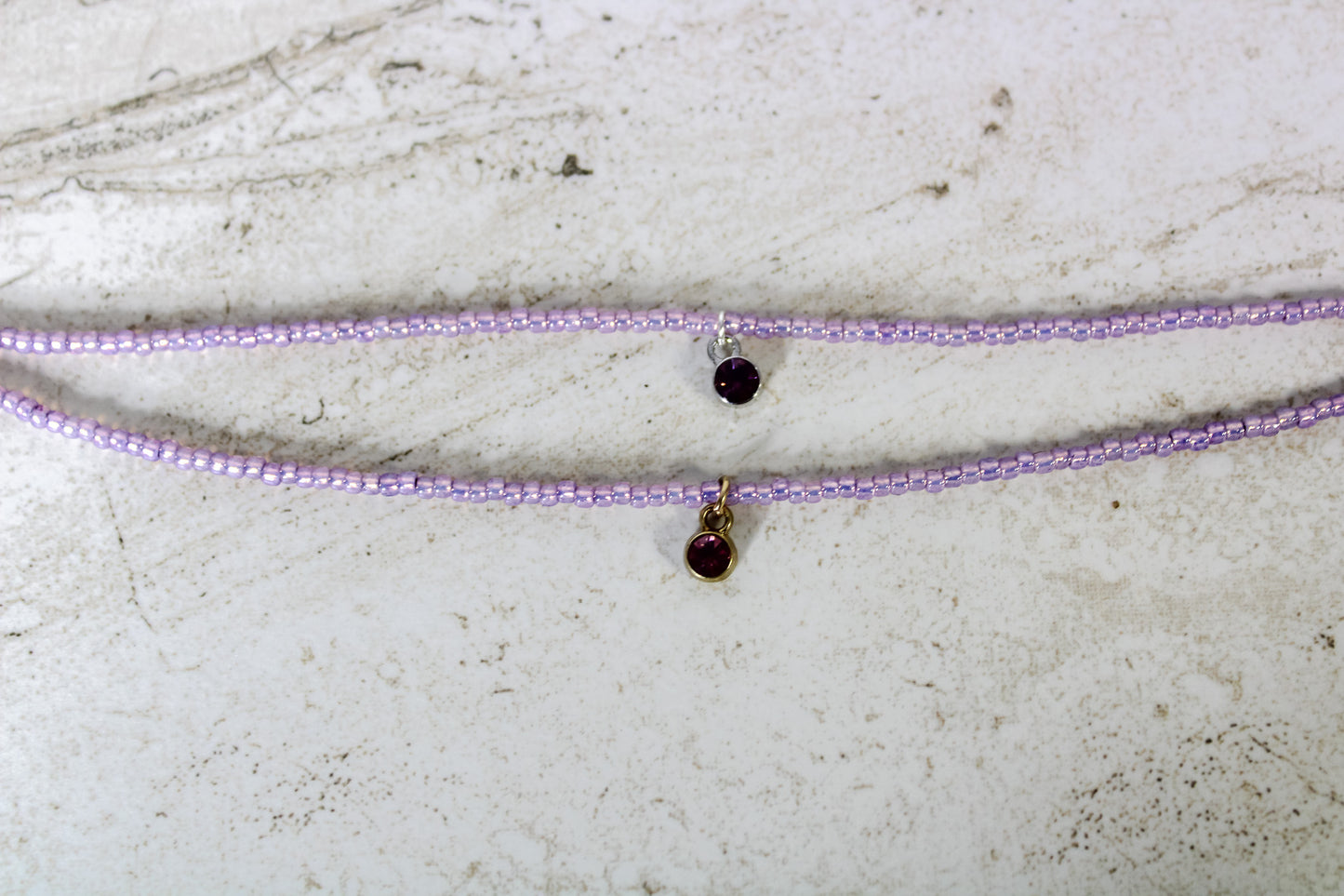 Two bracelets made out of purple seed beads and either a gold or silver circular amethyst cubic zirconia charm.