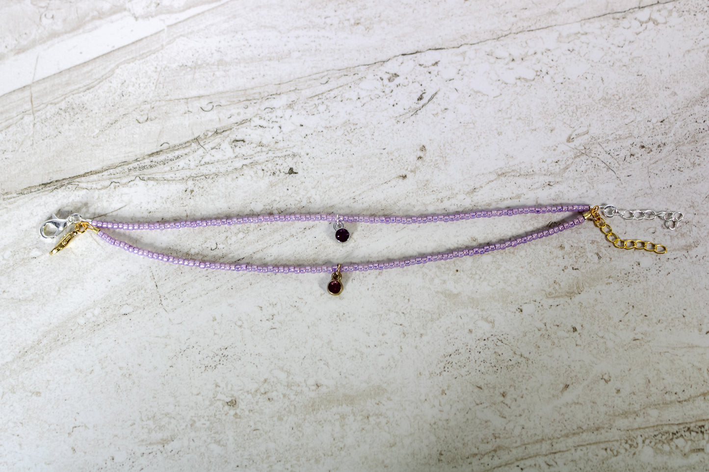 Two bracelets made out of purple seed beads and either a gold or silver circular amethyst cubic zirconia charm.