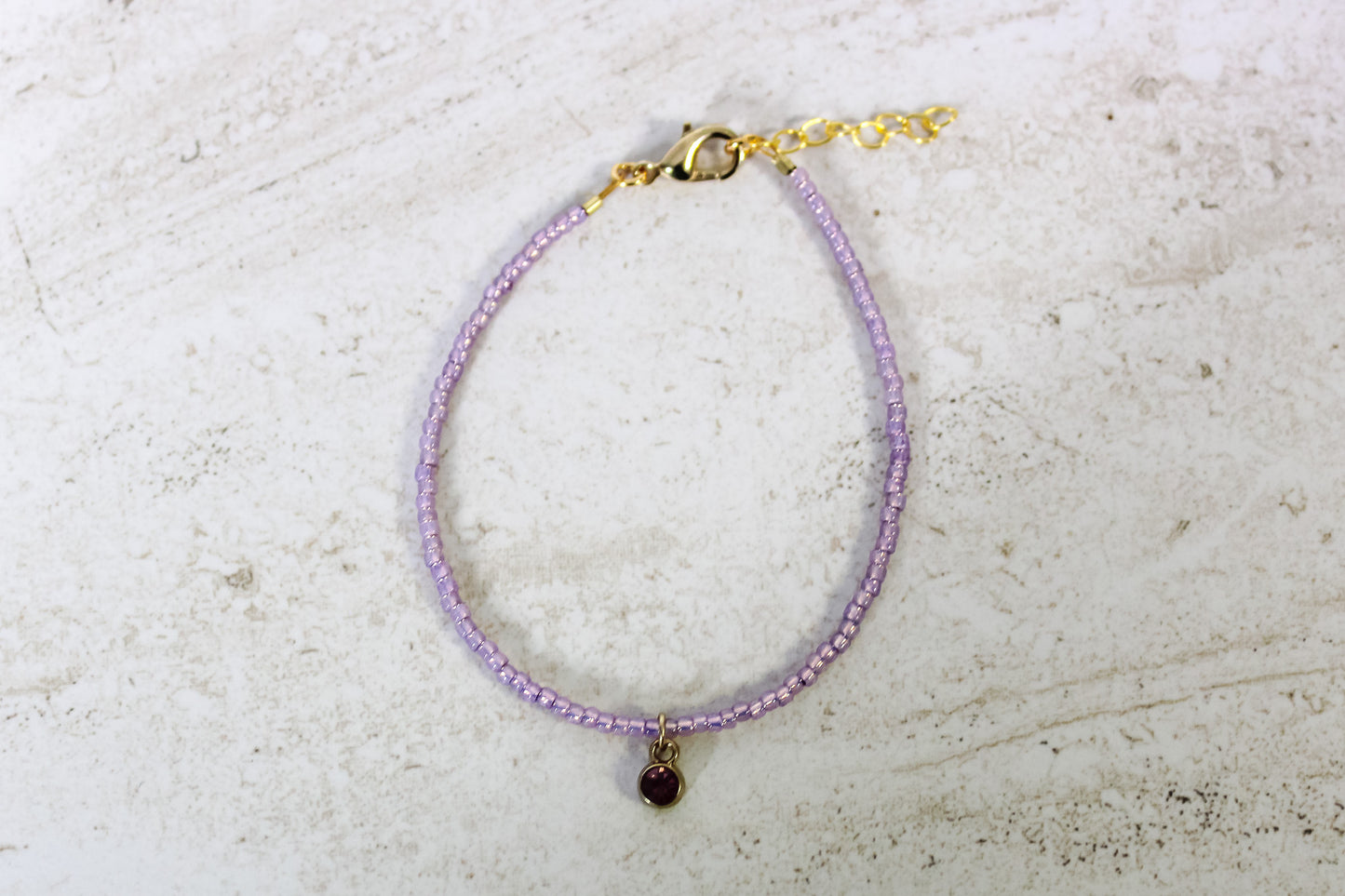 A purple colored seed bead bracelet with a small amethyst gold finished charm and gold lobster clasp and extender chain.