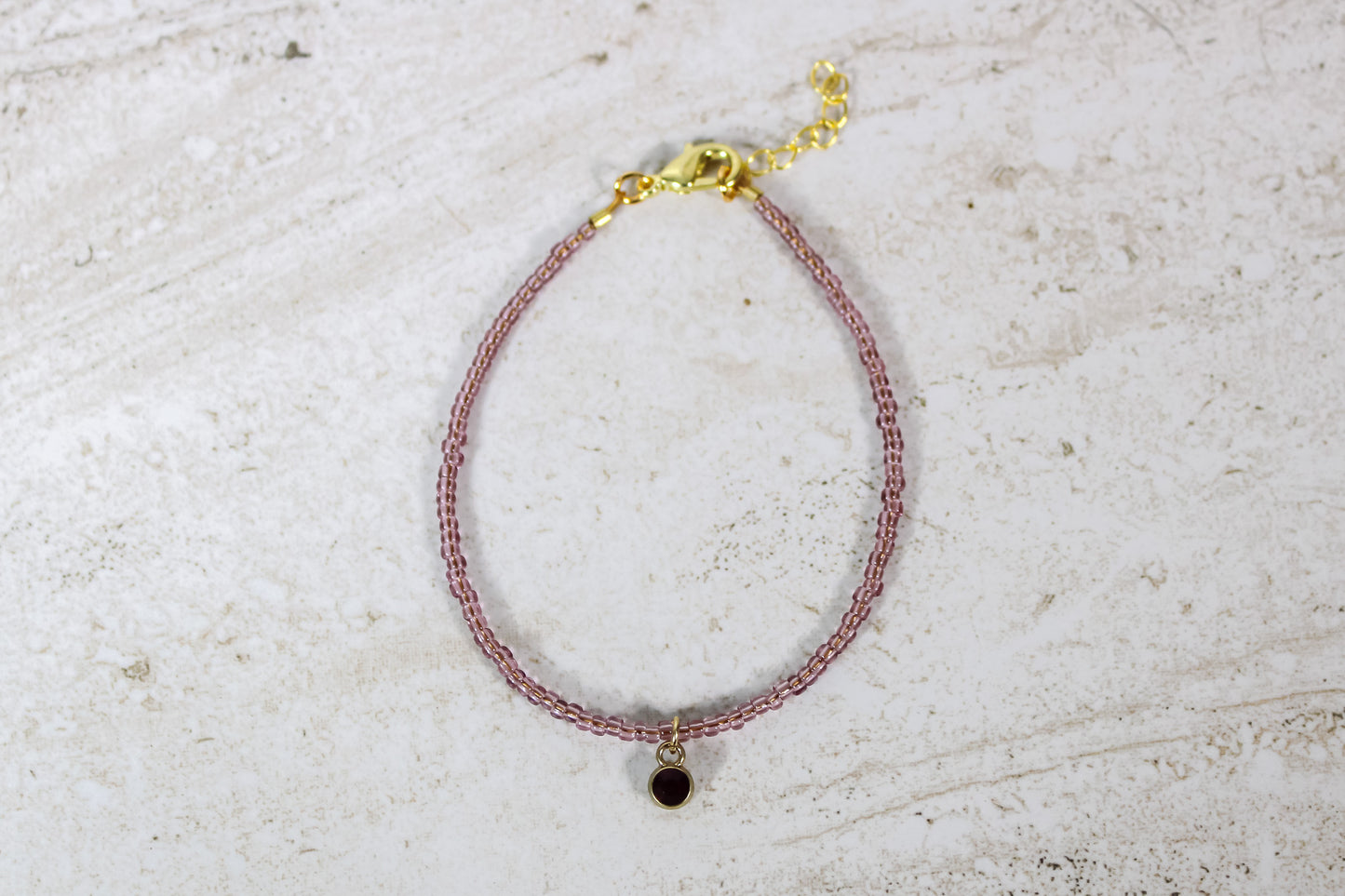 A garnet colored seed bead bracelet with a small garnet gold finished charm and gold lobster clasp and extender chain.