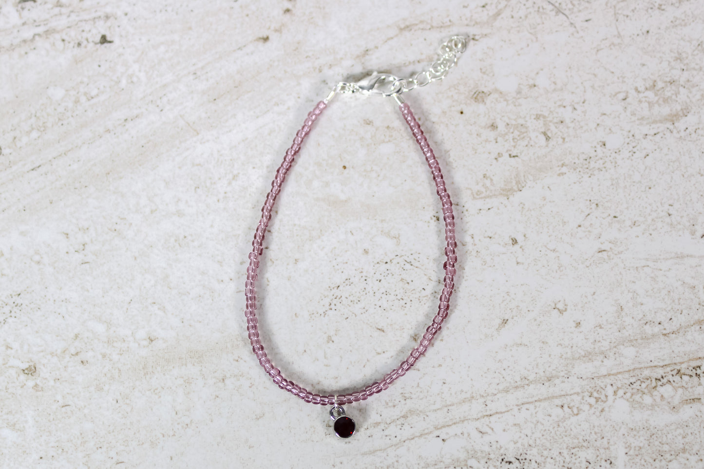 A garnet colored seed bead bracelet with a small garnet silver finished charm and silver lobster clasp and extender chain.