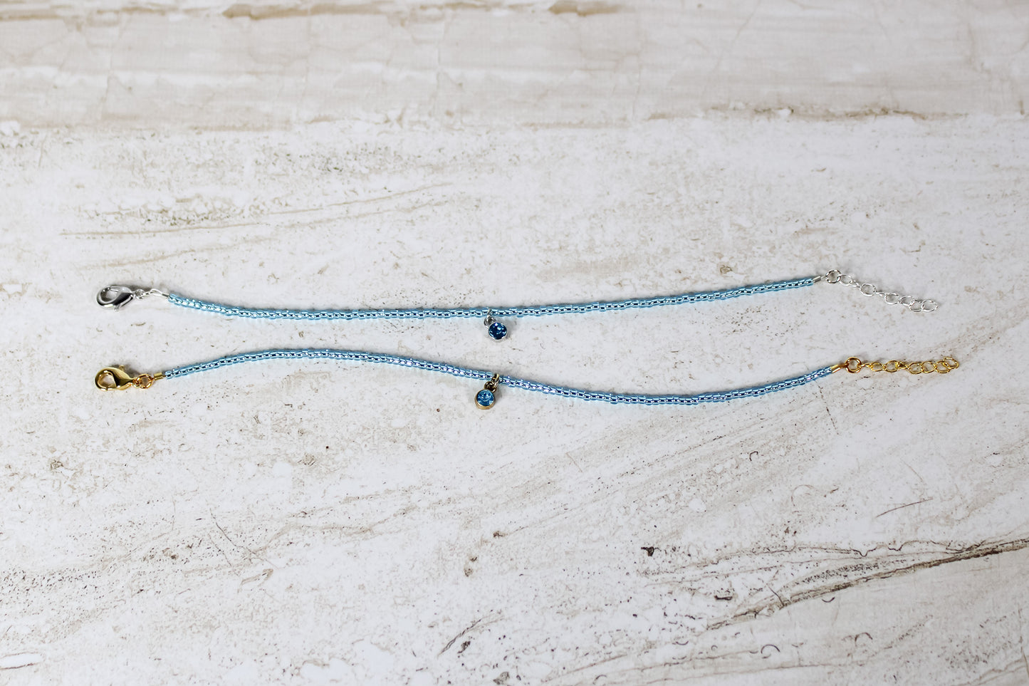 Two bracelets made out of aquamarine seed beads and either a gold or silver circular aquamarine cubic zirconia charm.