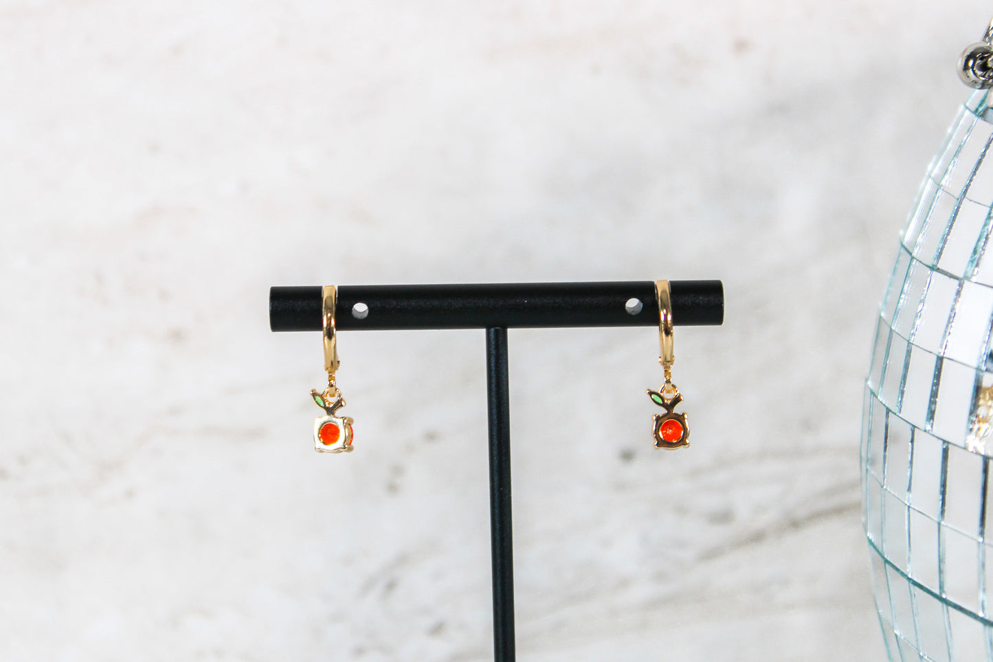 A pair of gold huggie hoop earrings with small orange fruit cubic zirconia charms are displayed on a t bar, showing the back of the earrings.