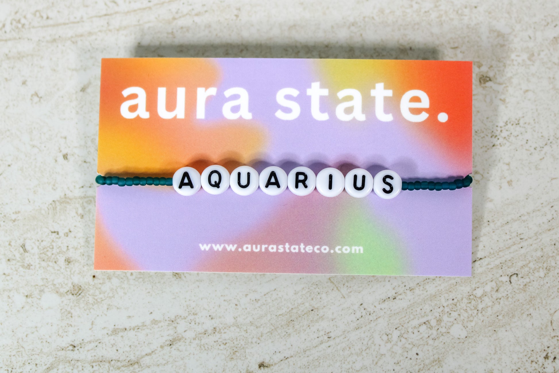 A teal beaded bracelet with plastic round beads that spell Aquarius on a rectangular card that says aura state.