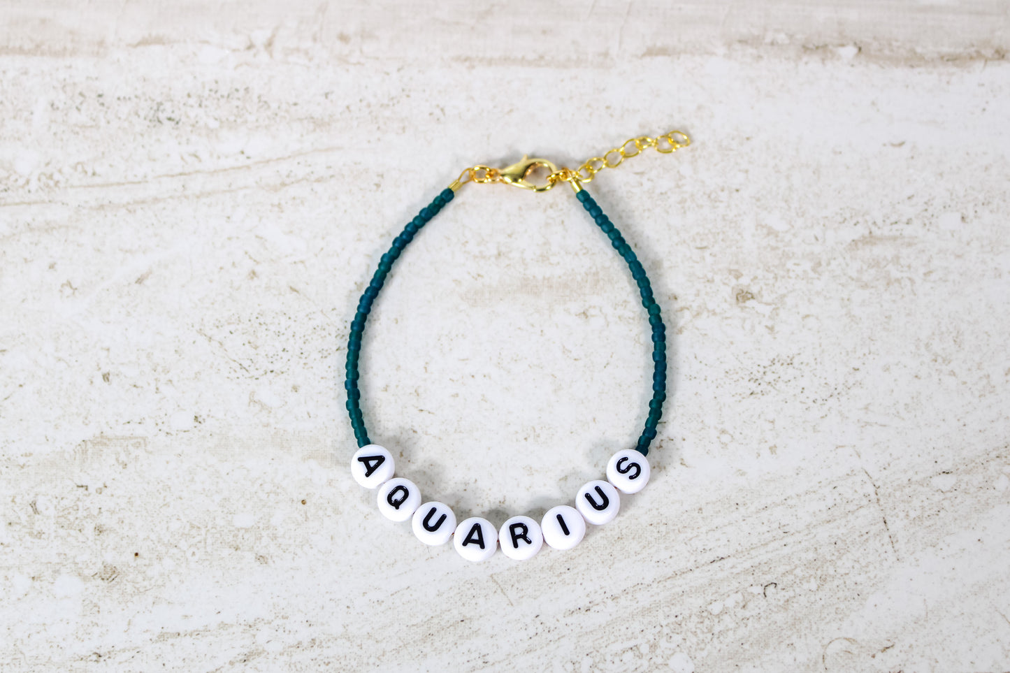 A teal colored seed bead bracelet with a Aquarius in round letter beads and gold lobster clasp and extender chain.