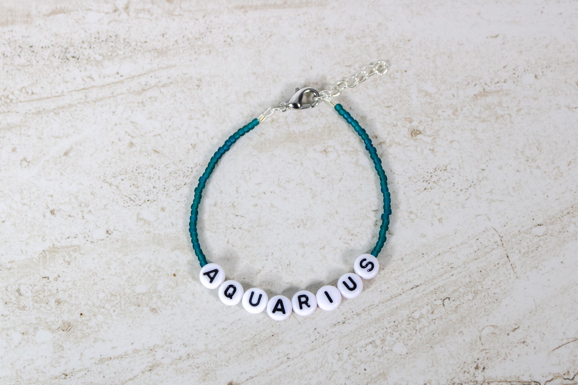 A teal colored seed bead bracelet with a Aquarius in round letter beads and silver lobster clasp and extender chain.