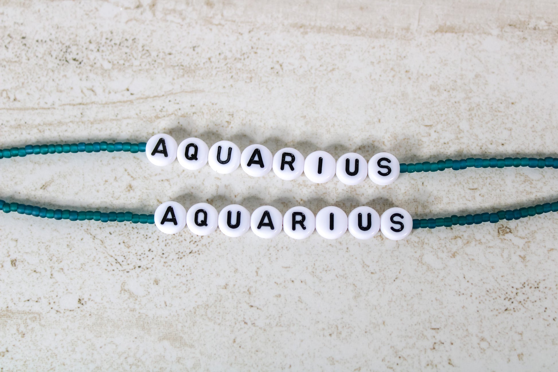 Two bracelets made out of teal seed beads with plastic beads that spell out Aquarius.