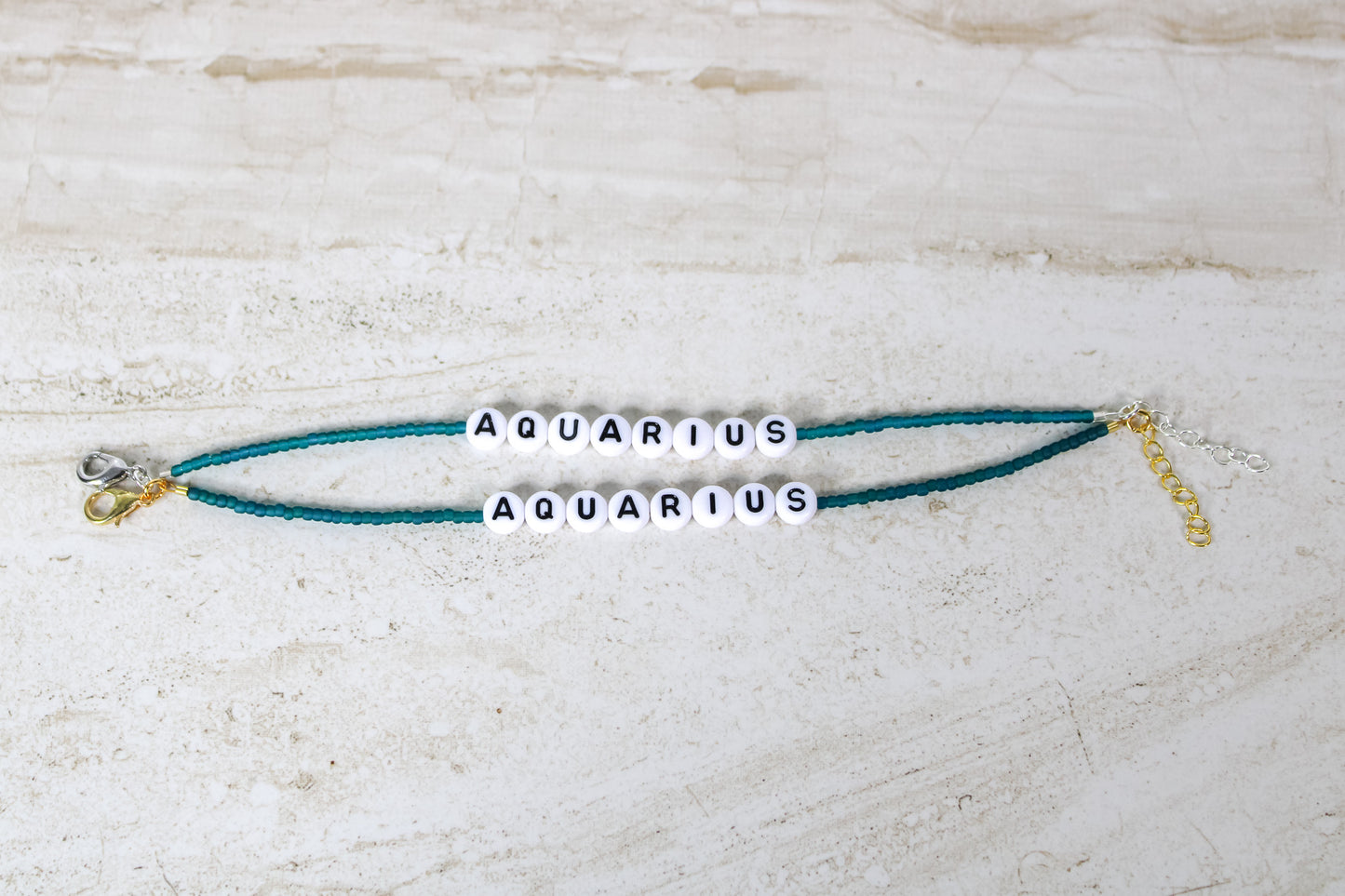 Two bracelets made out of teal seed beads with plastic beads that spell out Aquarius.