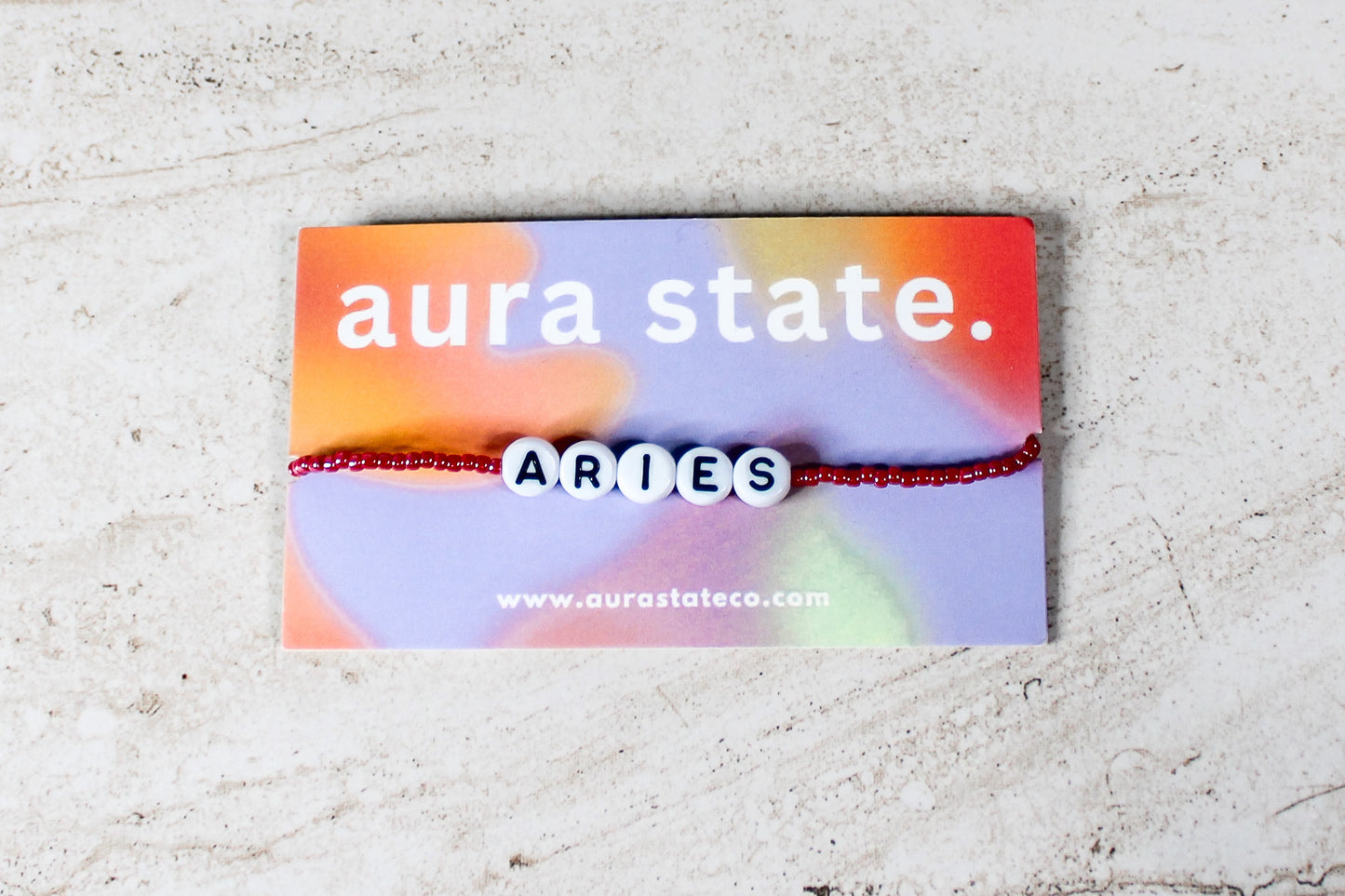 A shiny beaded bracelet with plastic round beads that spell Aries on a rectangular card that says aura state.