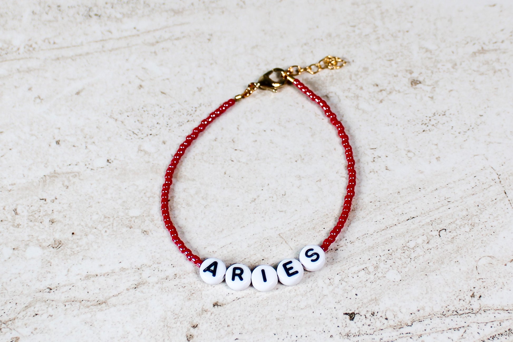 A shiny red colored seed bead bracelet with Aries in round letter beads and gold lobster clasp and extender chain.