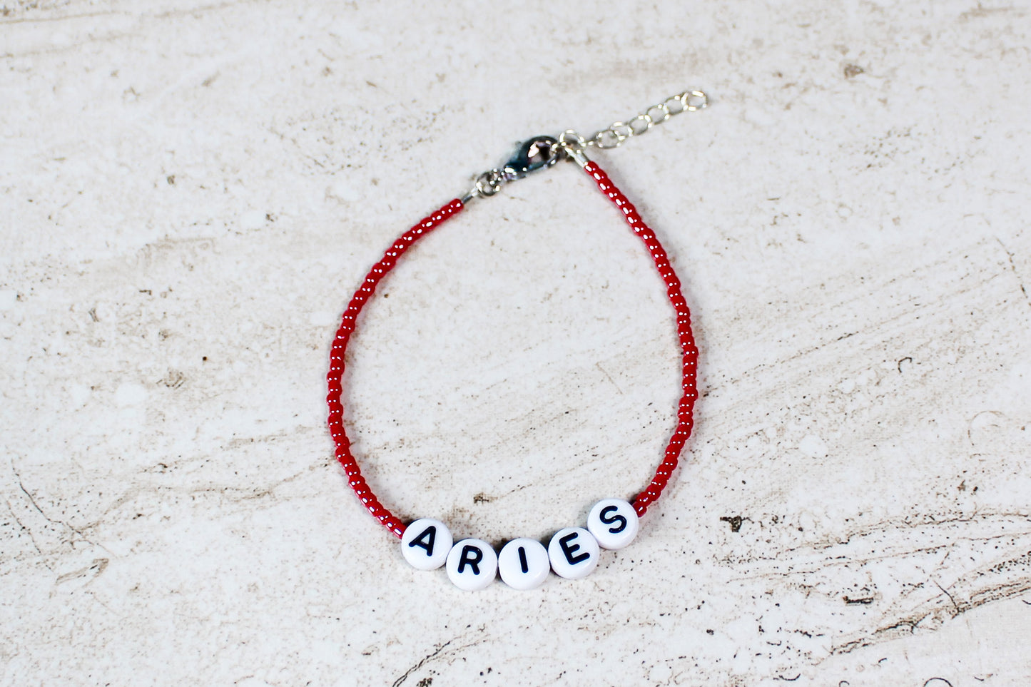 A shiny red colored seed bead bracelet with Aries in round letter beads and silver lobster clasp and extender chain.