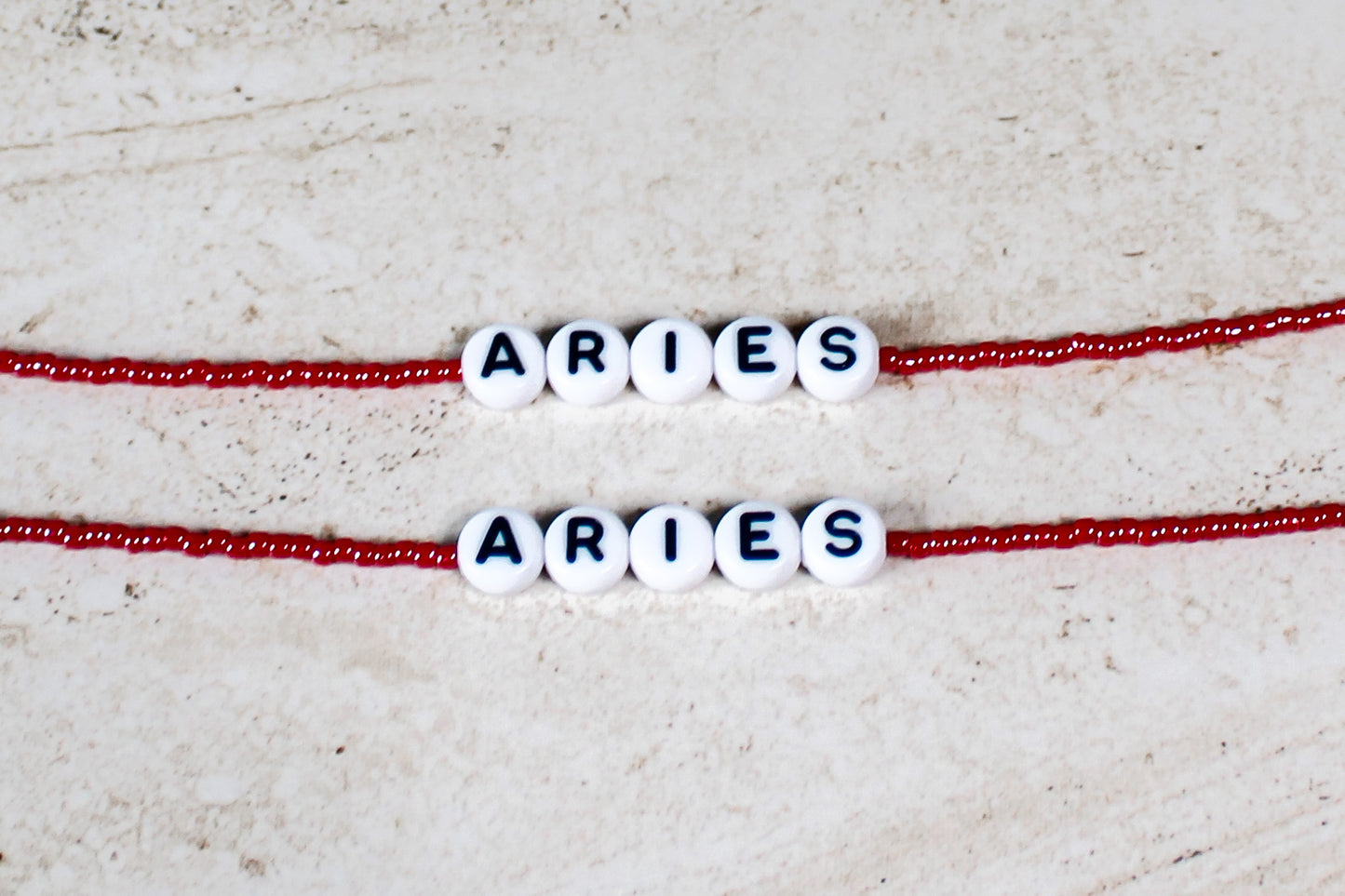 Two bracelets made out of shiny red seed beads with plastic beads that spell out Aries.