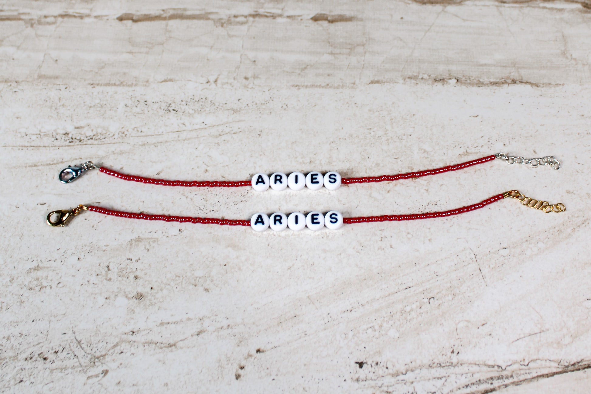 Two bracelets made out of shiny red seed beads with plastic beads that spell out Aries.