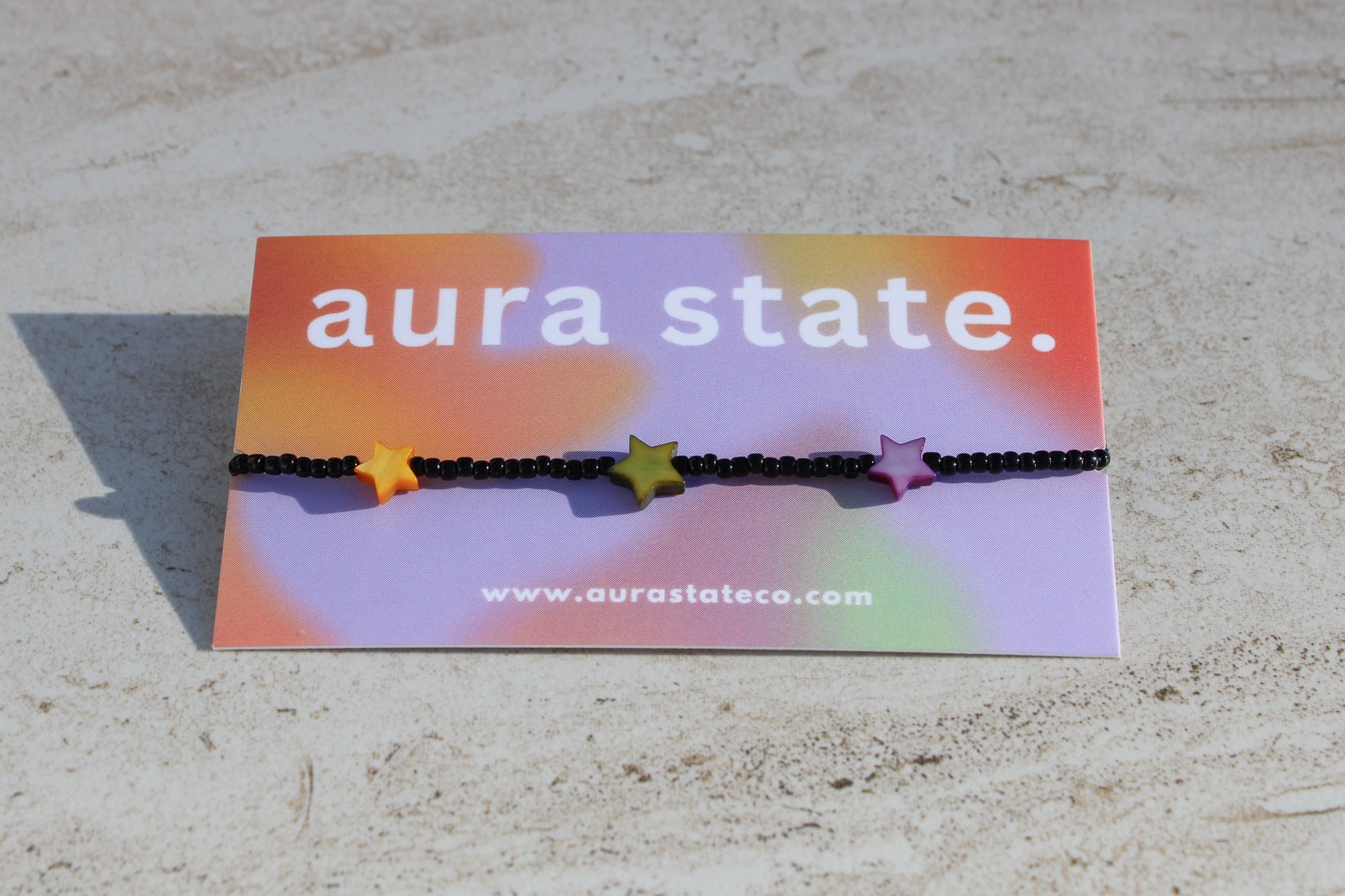 A black beaded bracelet with yellow, green, and purple shell star charms on a rectangular card that says aura state.