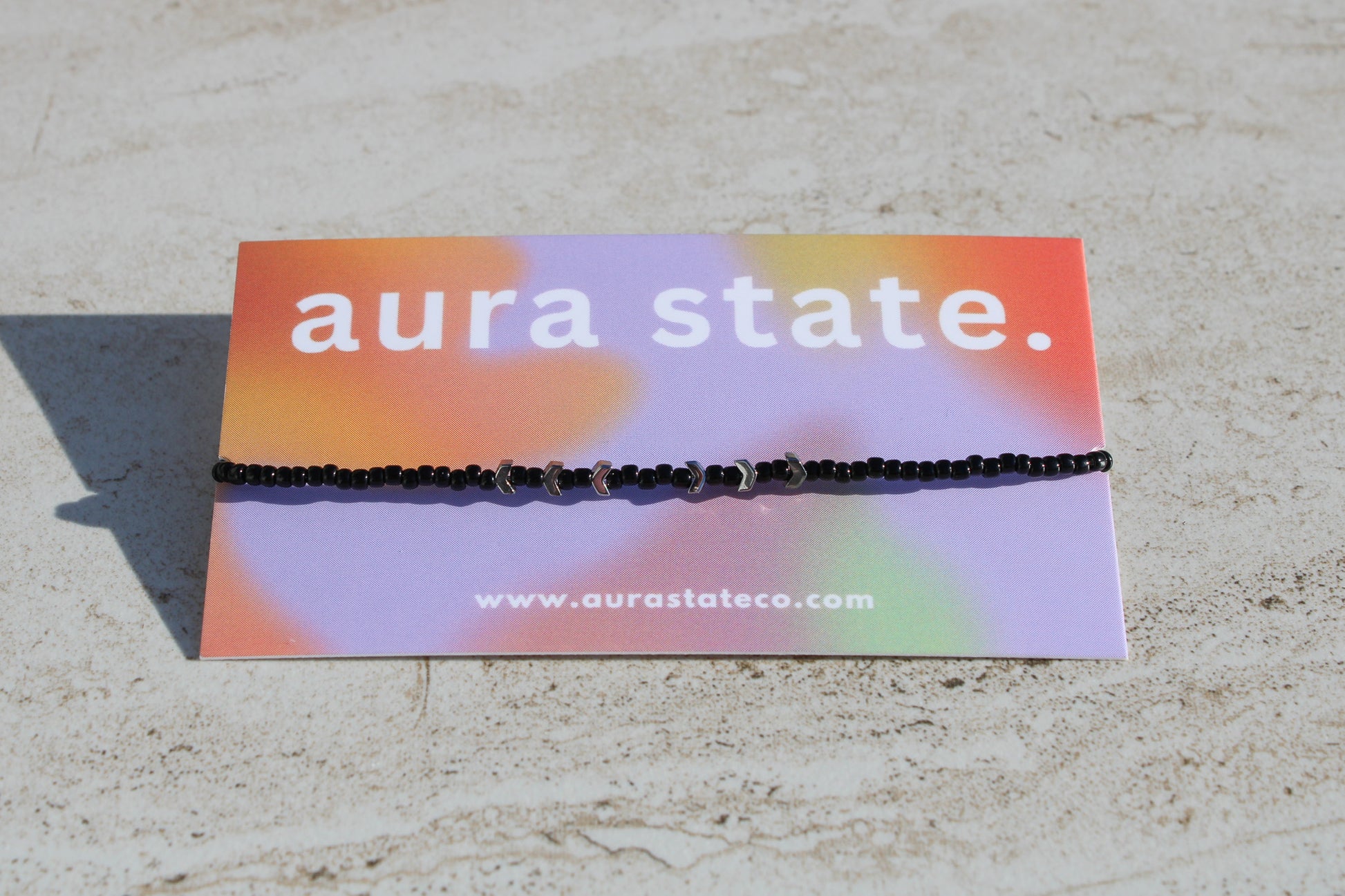 A black beaded bracelet with silver arrow charms on a rectangular card that says aura state.