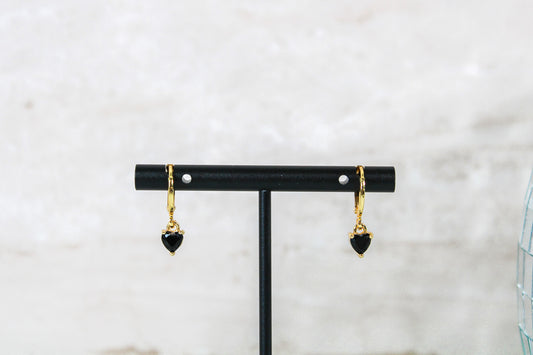 A pair of gold huggie hoop earrings with small black cubic zirconia heart charms are displayed on a t bar.