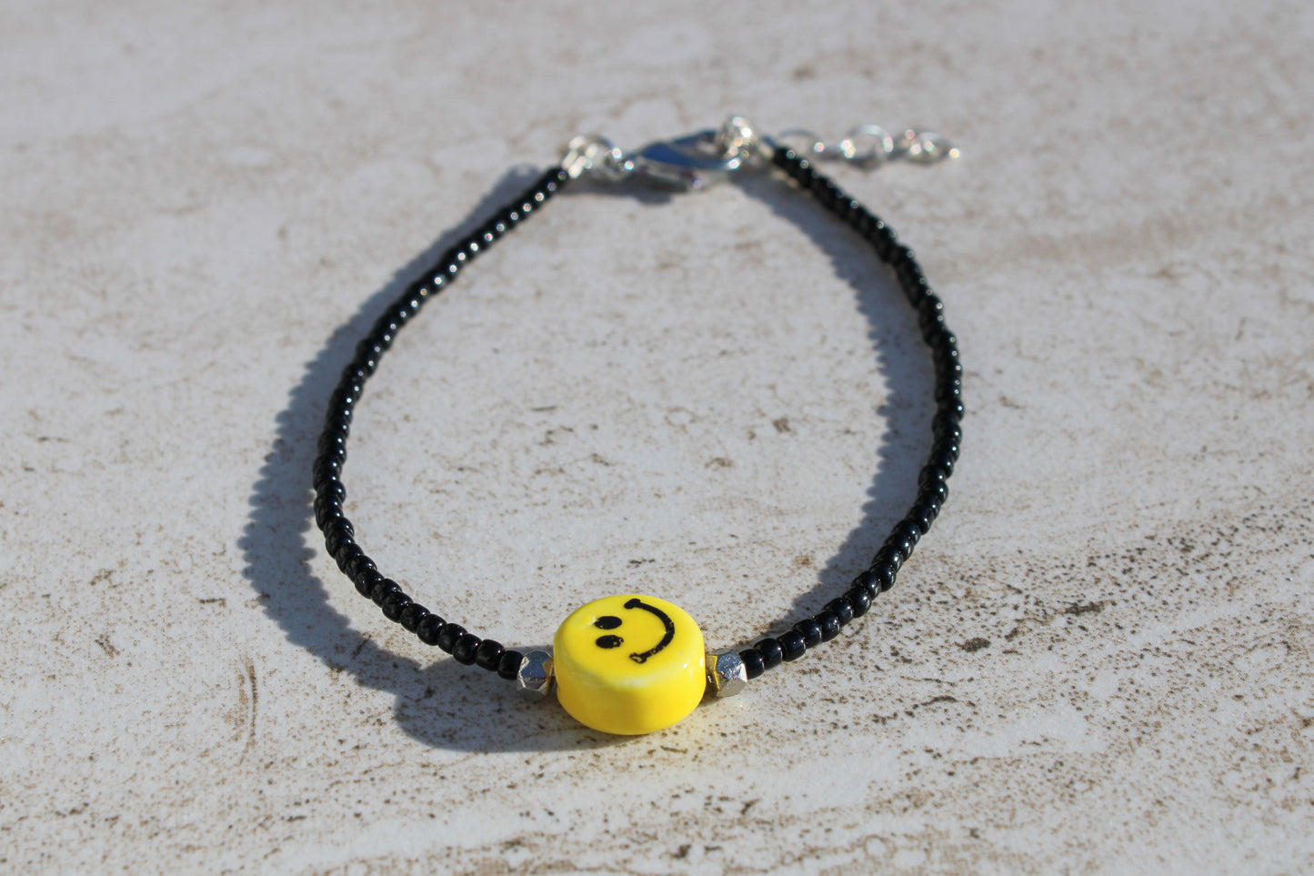 A bracelet made out of black seed beads and a yellow smiley face charm in the middle supported by two small silver beads on either side.  Features a silver lobster claw clasp and chain extender.