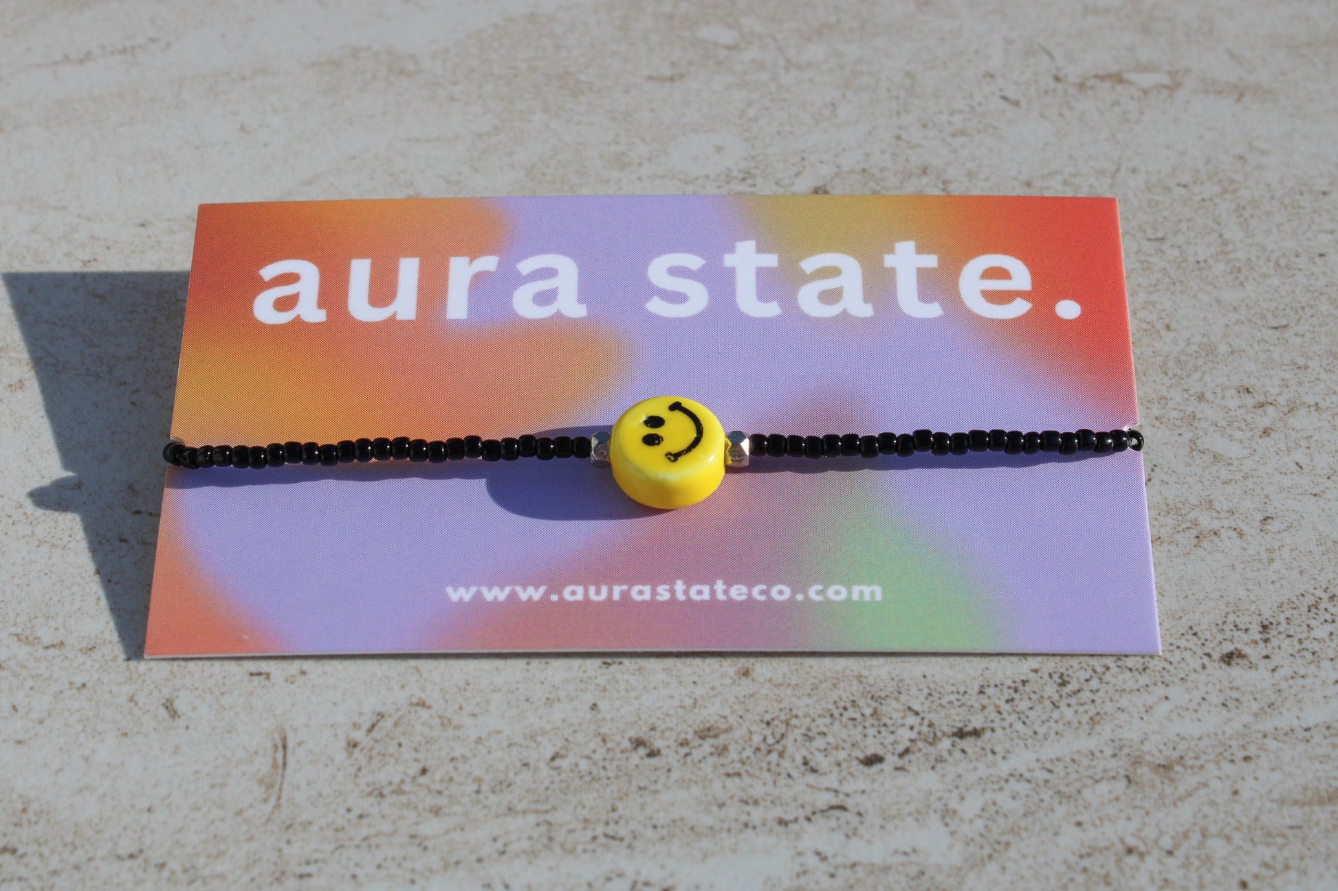 A black beaded bracelet with a yellow smiley face charm on a rectangular card that says aura state.