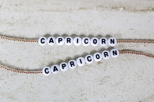 Two bracelets made out of copper seed beads with plastic beads that spell out Capricorn.