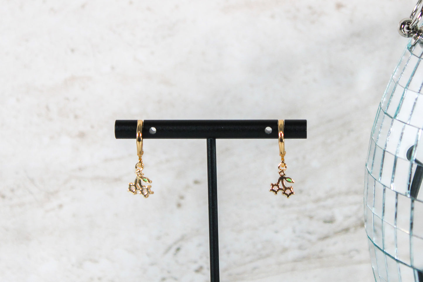 A pair of gold huggie hoop earrings with small cherry cubic zirconia charms are displayed on a t bar, showing the back of the earrings.