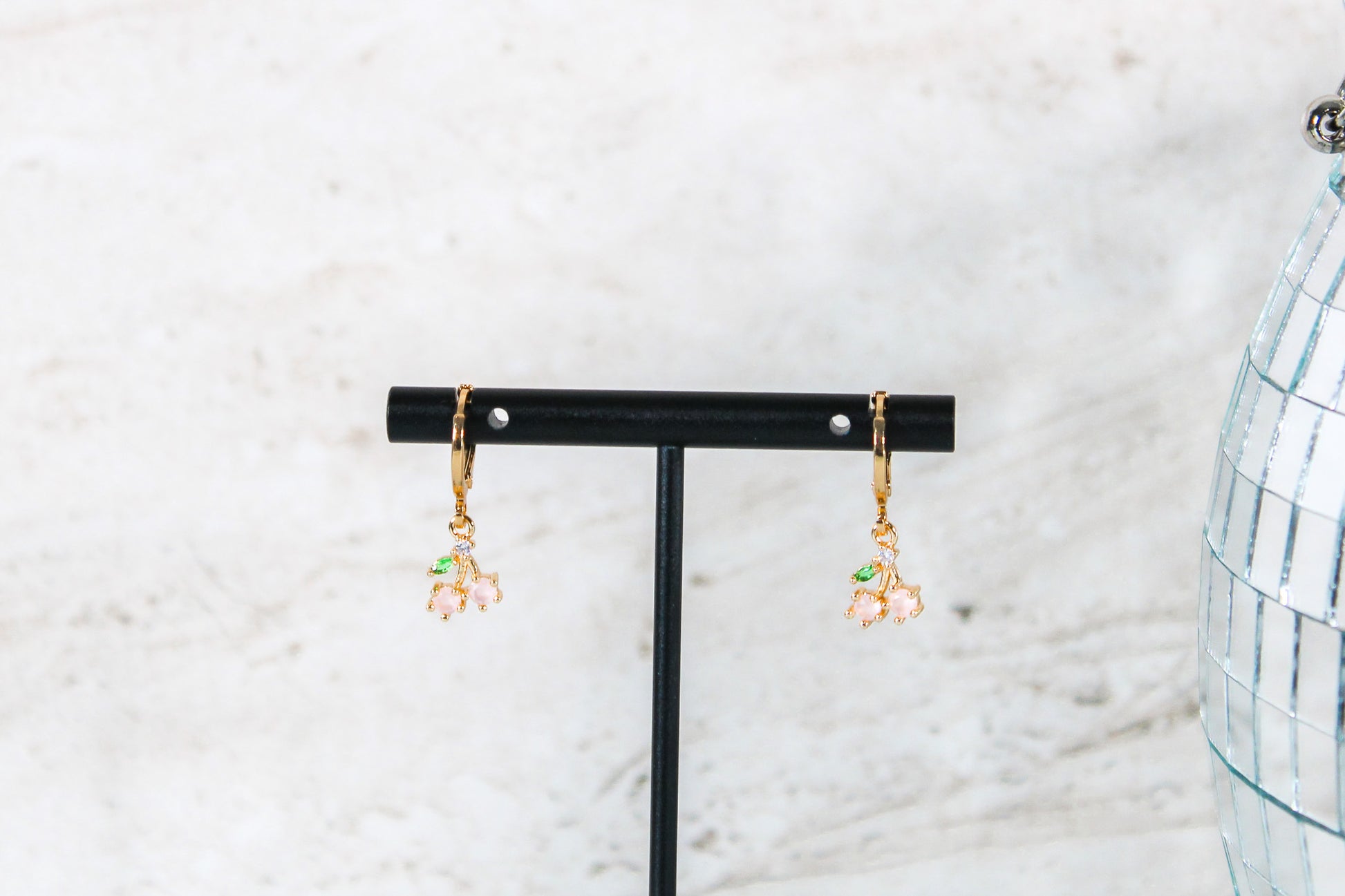 A pair of gold huggie hoop earrings with small cherry cubic zirconia charms are displayed on a t bar.