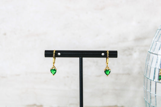 A pair of gold huggie hoop earrings with small green cubic zirconia heart charms are displayed on a t bar.