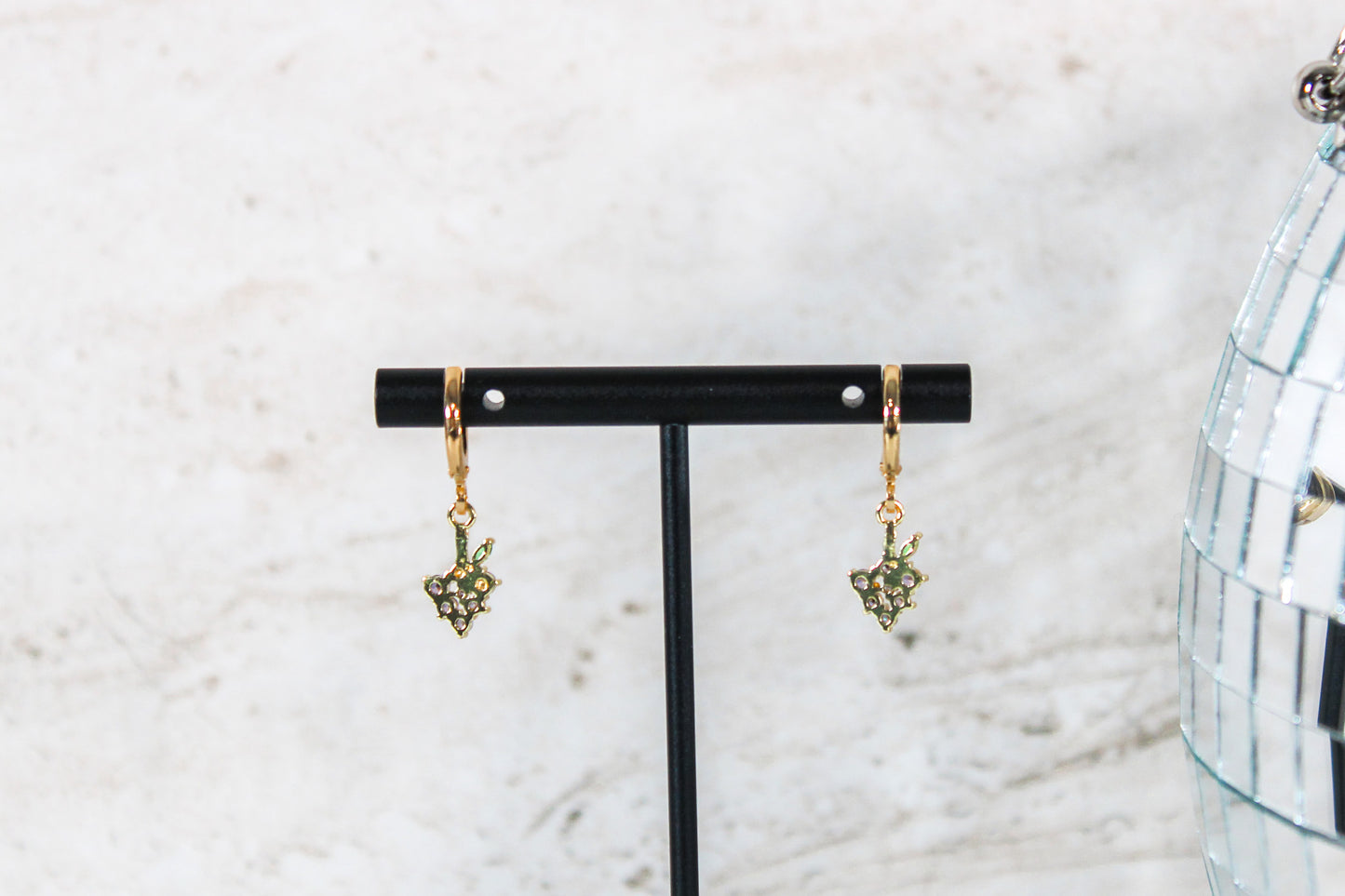 A pair of gold huggie hoop earrings with small grape cubic zirconia charms are displayed on a t bar, showing the back of the earrings.