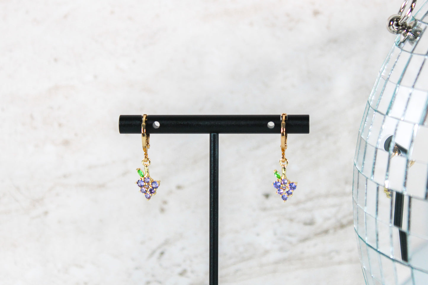 A pair of gold huggie hoop earrings with small grape cubic zirconia charms are displayed on a t bar.