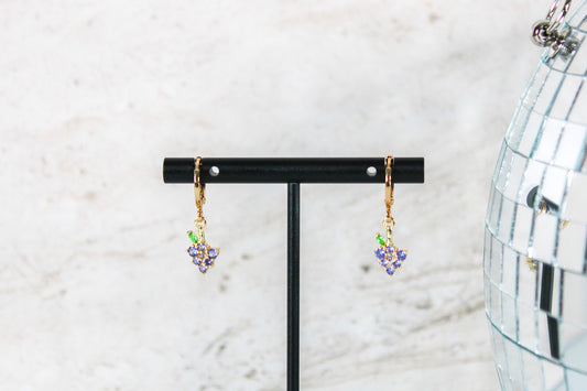 A pair of gold huggie hoop earrings with small grape cubic zirconia charms are displayed on a t bar.