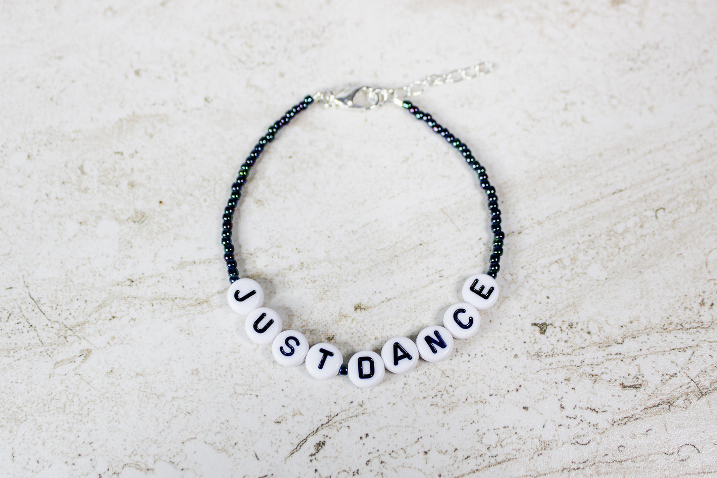 Handmade beaded bracelet that says Just Dance with oil slick seed beads and silver finishes, displayed in a flat lay view.