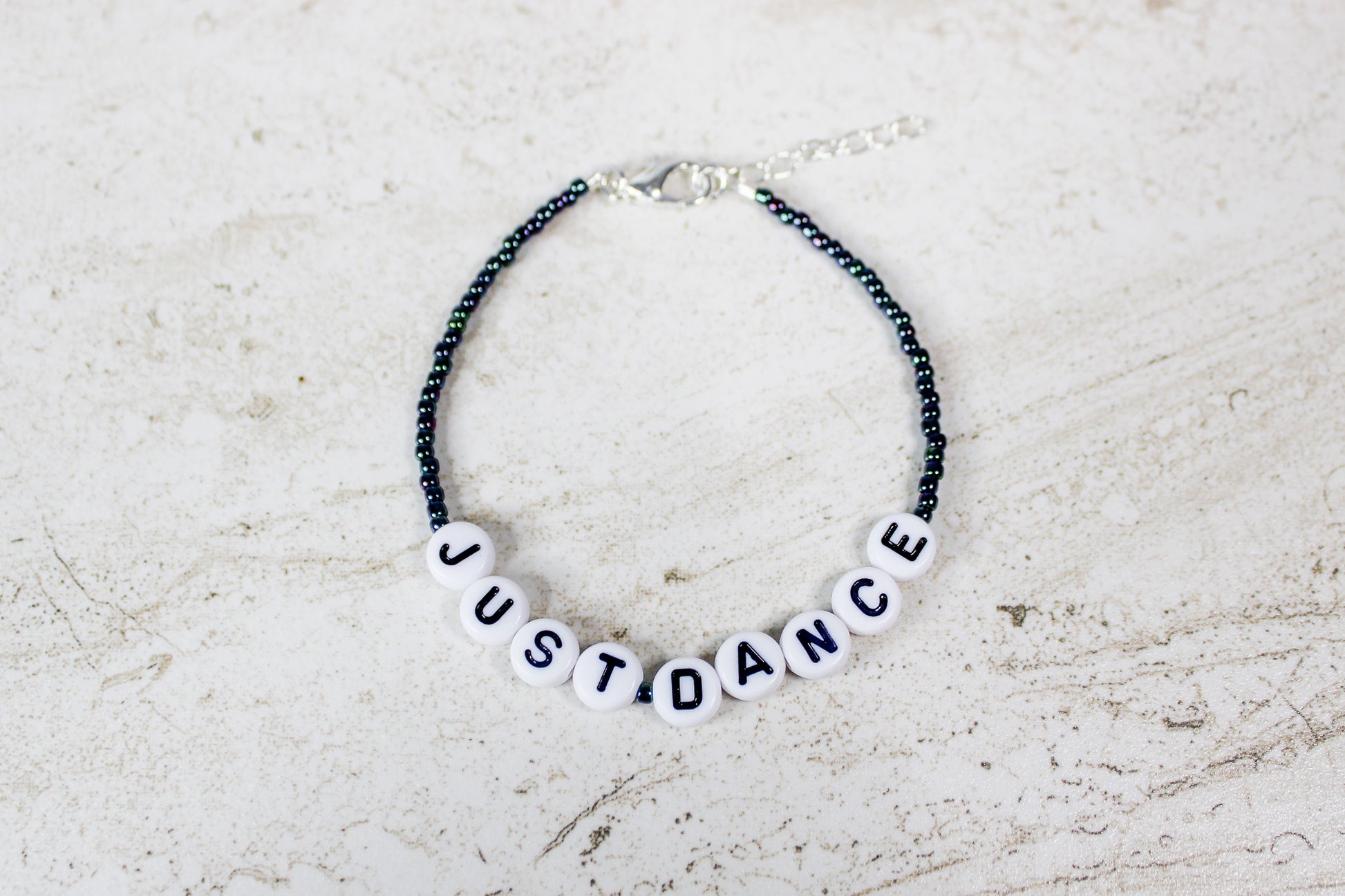 Handmade beaded bracelet that says Just Dance with oil slick seed beads and silver finishes, displayed in a flat lay view.