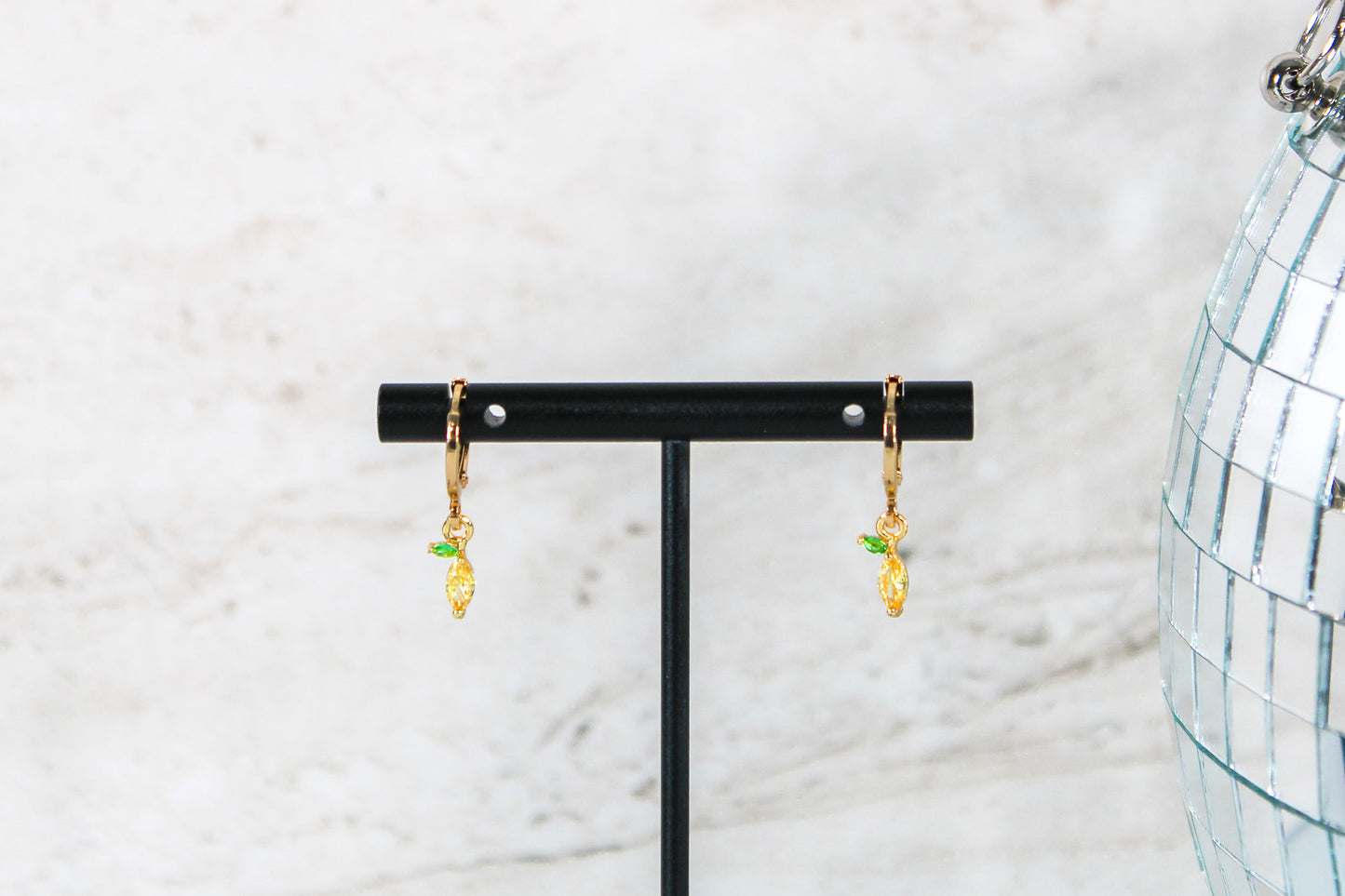 A pair of gold huggie hoop earrings with small lemon cubic zirconia charms are displayed on a t bar.