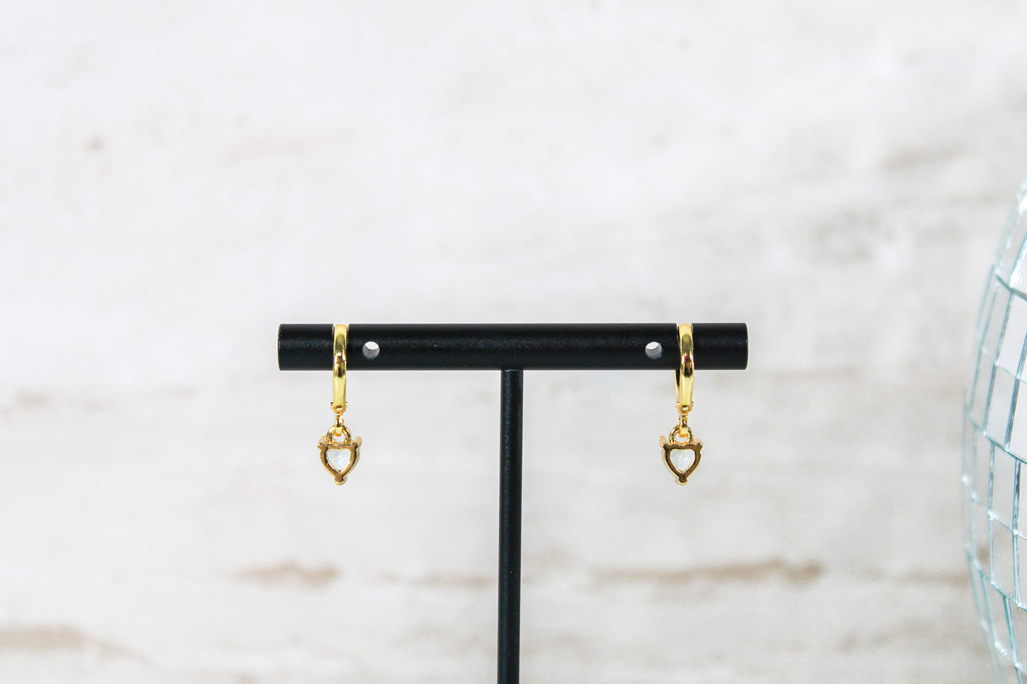 A pair of gold huggie hoop earrings with small blue cubic zirconia heart charms are displayed on a t bar, showing the back of the earrings.