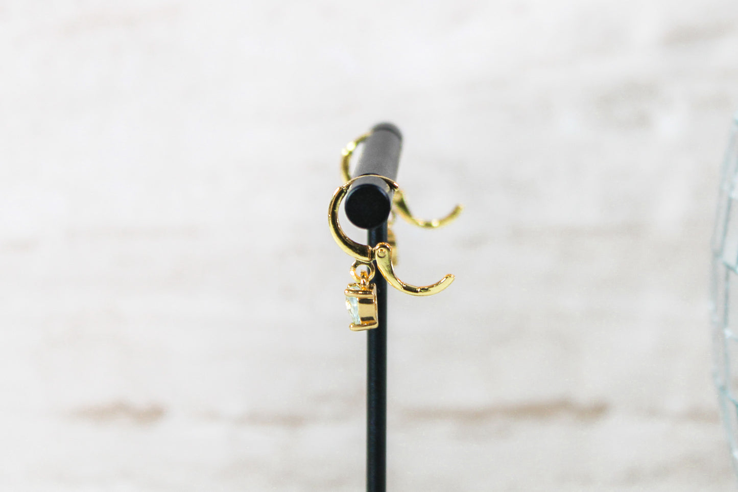 A pair of gold huggie hoop earrings with small blue cubic zirconia heart charms are displayed on a t bar, showing the open clasp of the earrings.