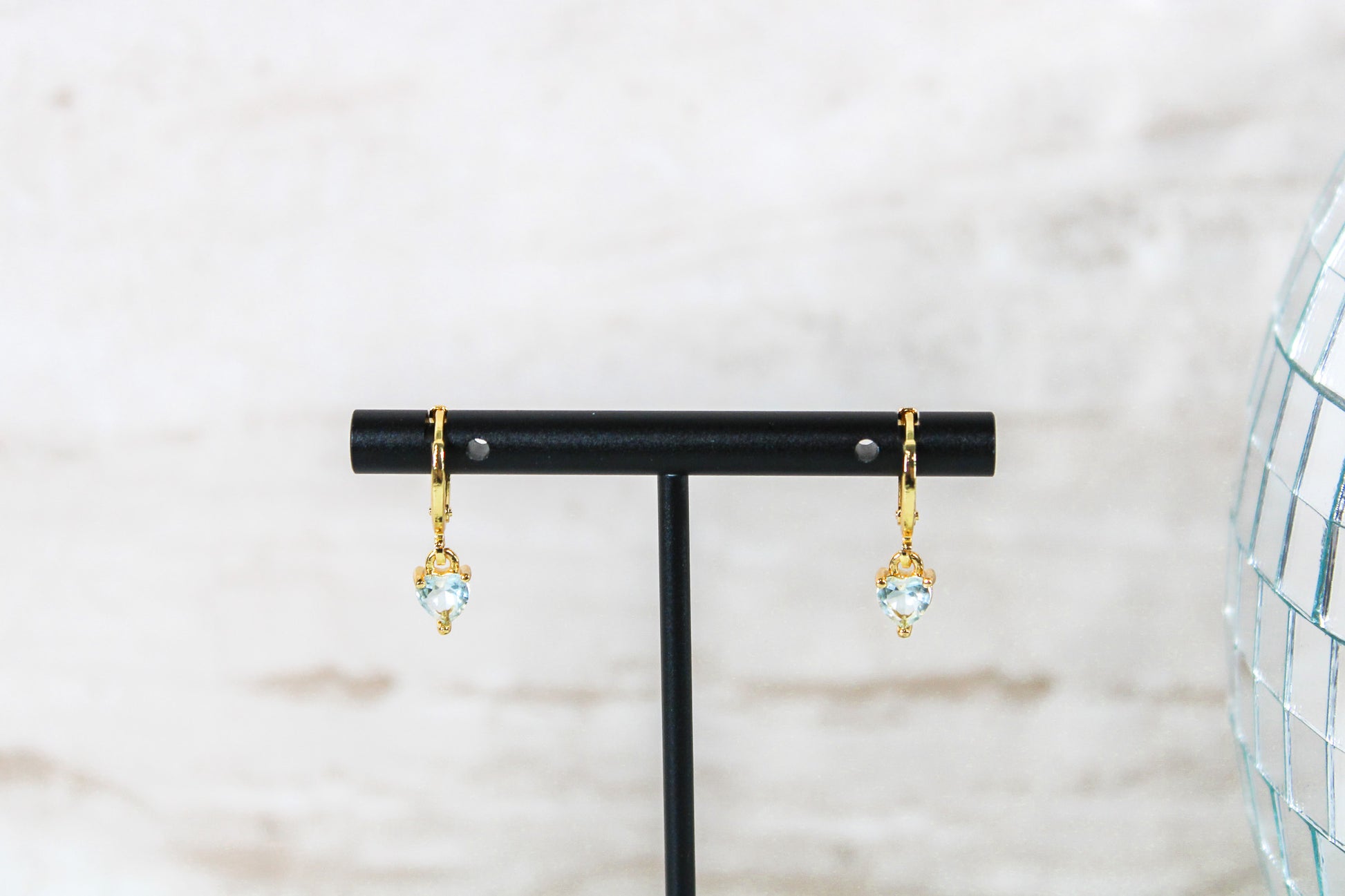 A pair of gold huggie hoop earrings with small blue cubic zirconia heart charms are displayed on a t bar.