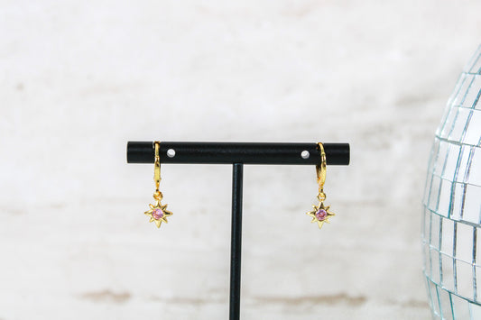 A pair of gold huggie hoop earrings with small pink cubic zirconia north star charms are displayed on a t bar.