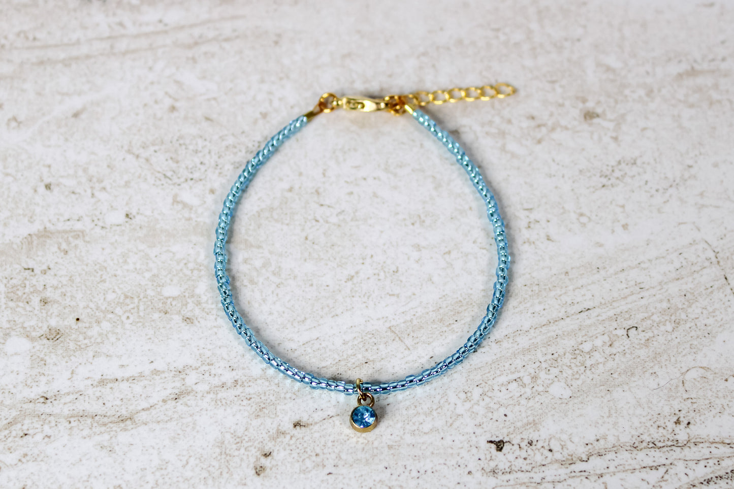 An aquamarine colored seed bead bracelet with a small aquamarine with gold finished charm and gold lobster clasp and extender chain.