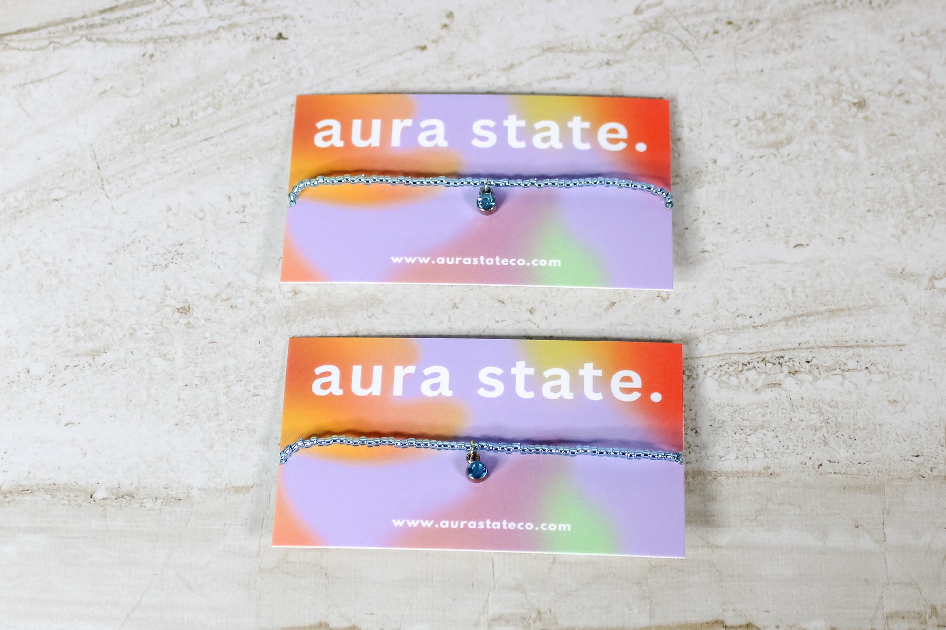 An aquamarine beaded bracelet with an aquamarine charm on a rectangular card that says aura state.