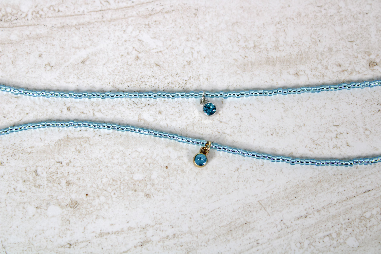 Two bracelets made out of aquamarine seed beads and either a gold or silver circular aquamarine cubic zirconia charm.