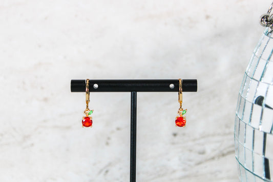 A pair of gold huggie hoop earrings with small orange fruit cubic zirconia charms are displayed on a t bar.