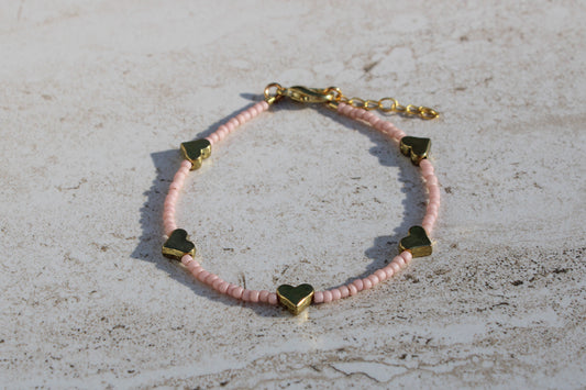 A bracelet made out of matte pale pink seed beads and 5 gold heart charms.  Features a gold lobster claw clasp and chain extender.