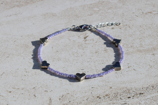 A bracelet made out of pastel purple seed beads and silver heart charms.  Features a silver lobster claw clasp and chain extender.