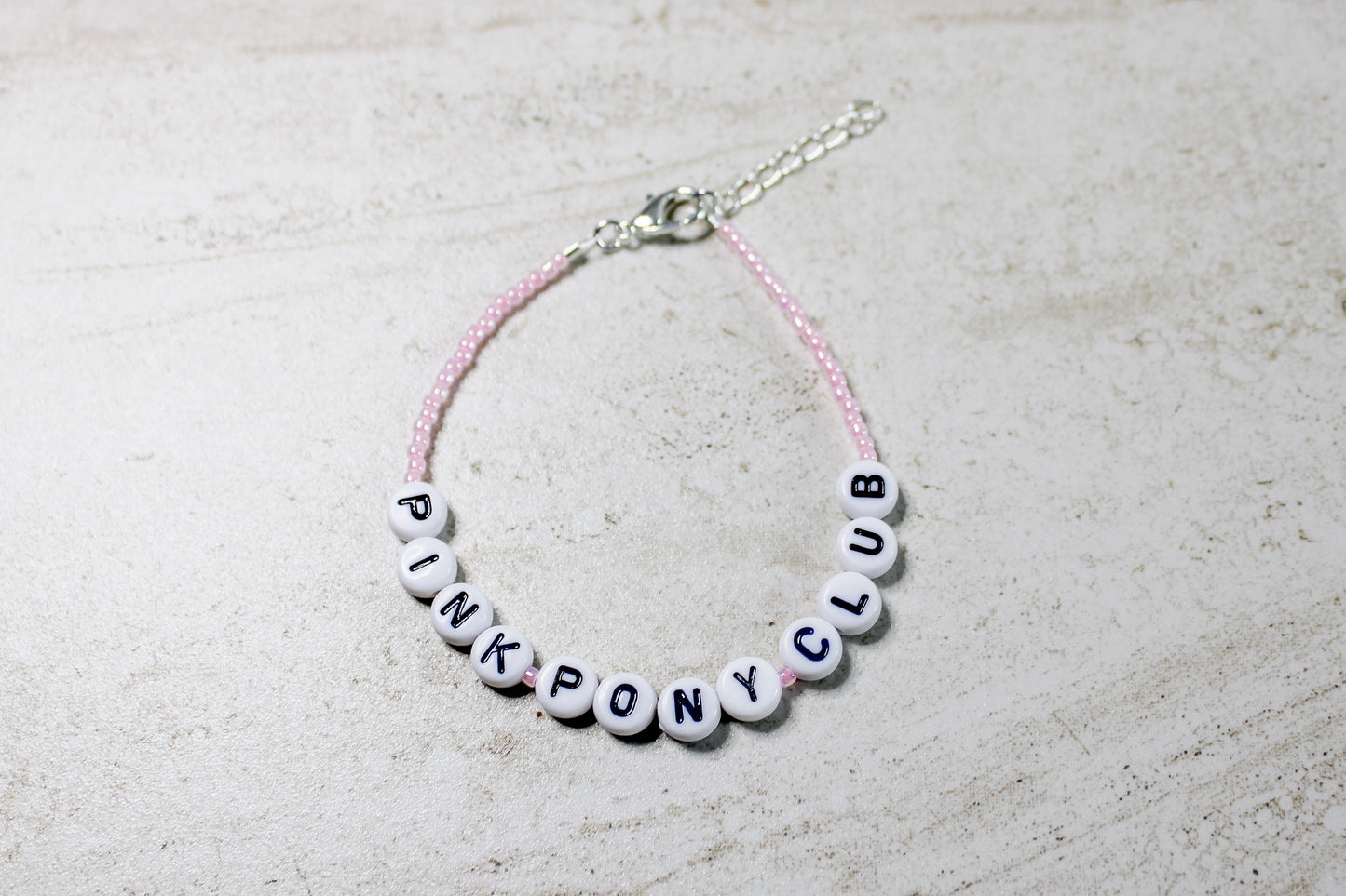 Light pink beaded bracelet with “Pink Pony Club” spelled in white letter beads, laid flat in a circular shape.