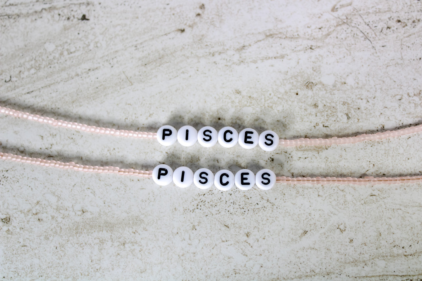 Two bracelets made out of pale pink seed beads with plastic beads that spell out Pisces.