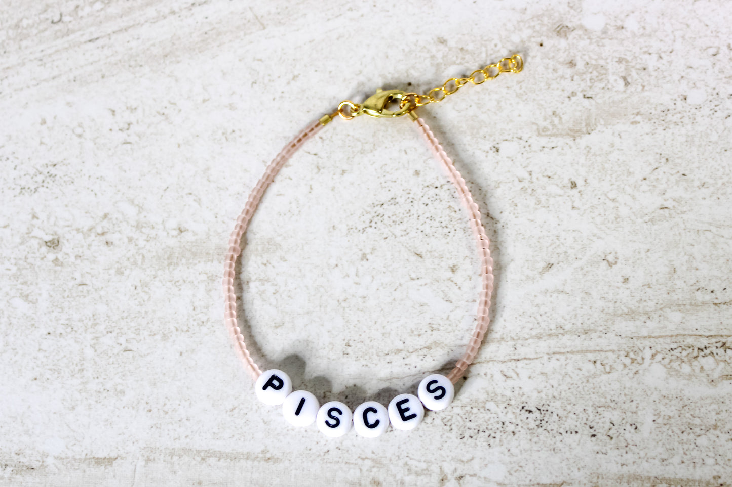 A pale pink colored seed bead bracelet with Pisces in round letter beads and gold lobster clasp and extender chain.