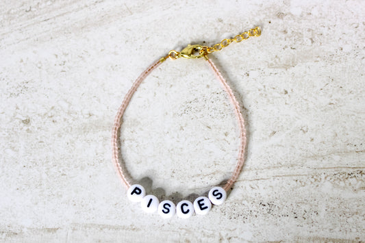 A pale pink colored seed bead bracelet with Pisces in round letter beads and gold lobster clasp and extender chain.