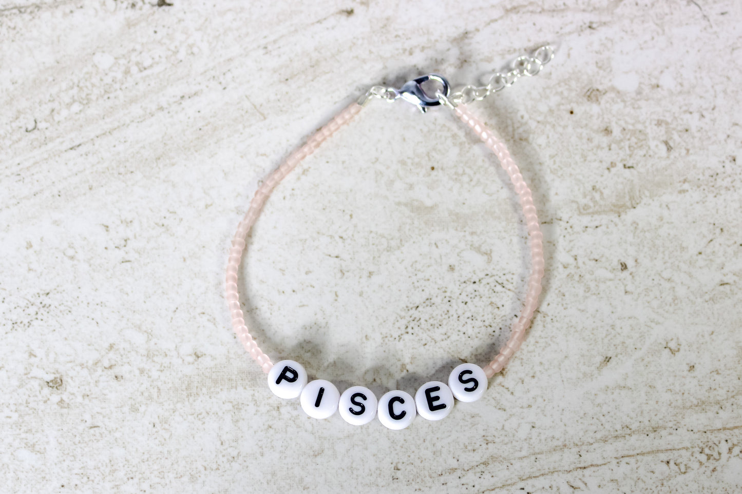 A pale pink colored seed bead bracelet with Pisces in round letter beads and silver lobster clasp and extender chain.